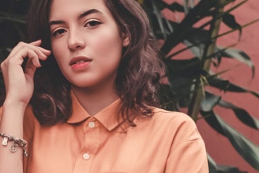 A fashionable woman with brown hair, wearing an orange blouse and a gold charm bracelet